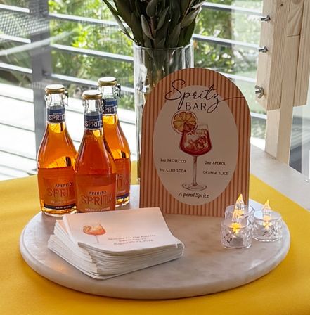 Spritz bar cocktail station with three orange aperitif spritz bottles, striped menu sign listing a spritz recipe, stacked napkins and glowing LED candles on a round marble board by a sunlit patio window.