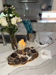 Stylish modern kitchen island with white marble countertop and gray subway tile backsplash, featuring a vase of white roses, three flameless candles, individually wrapped chocolate cookies on a gold tray, teal rolled items in a metal holder, and stacked cocktail napkins.