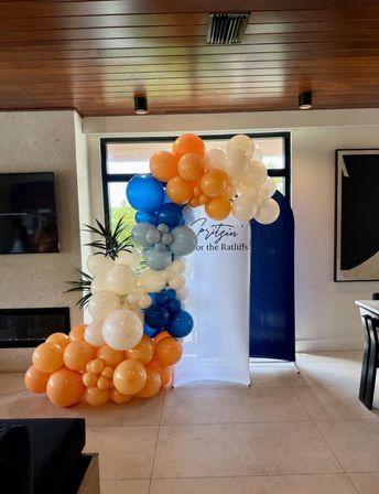 Cheerful indoor balloon garland in a modern living room: clustered orange, cream, blue and pale gray balloons framing a white-and-navy photo backdrop beside a window and plant.