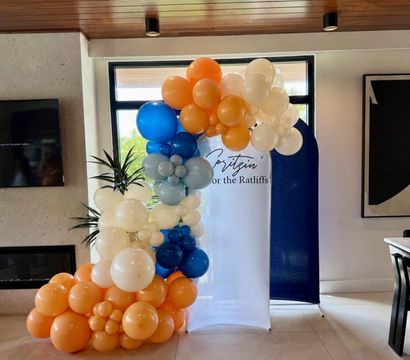Cheerful indoor balloon garland in a modern living room: clustered orange, cream, blue and pale gray balloons framing a white-and-navy photo backdrop beside a window and plant.