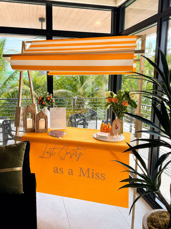 Bright orange-and-white striped canopy above a tropical balcony drink cart draped with an orange cloth reading "Last Spritz as a Miss", styled with fresh flowers, lanterns and aperitif bottles against glass windows with palm trees outside.