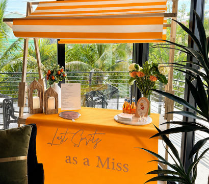 Bright orange-and-white striped canopy above a tropical balcony drink cart draped with an orange cloth reading "Last Spritz as a Miss", styled with fresh flowers, lanterns and aperitif bottles against glass windows with palm trees outside.