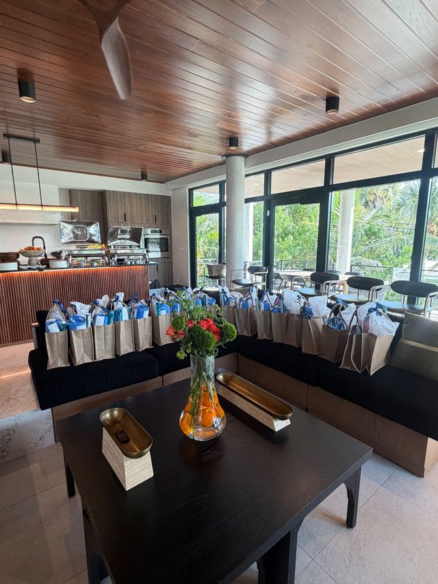 Sunlit modern villa living area with wooden ceiling and floor-to-ceiling windows, buffet kitchen, row of reusable gift bags on a black sectional, and a flower-filled vase on the coffee table with tropical greenery visible outside.