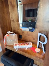 Bridal welcome on a wooden vanity: 'BRIDE' gift bag with a colorful scarf, ribbon-tied box of chocolates, oversized letter R, and a mirror reflecting a modern bedroom with TV and dresser.