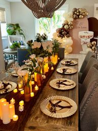 Indoor romantic dining room tablescape with a rustic wood table, white roses in amber vases, flickering LED candles, gold charger plates and a beige-and-gold balloon arch backdrop.