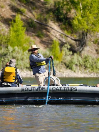 River rafting guide steering an inflatable raft with a long oar, passengers in bright life jackets and leafy riverbanks in the background — sunny outdoor adventure on a scenic river.