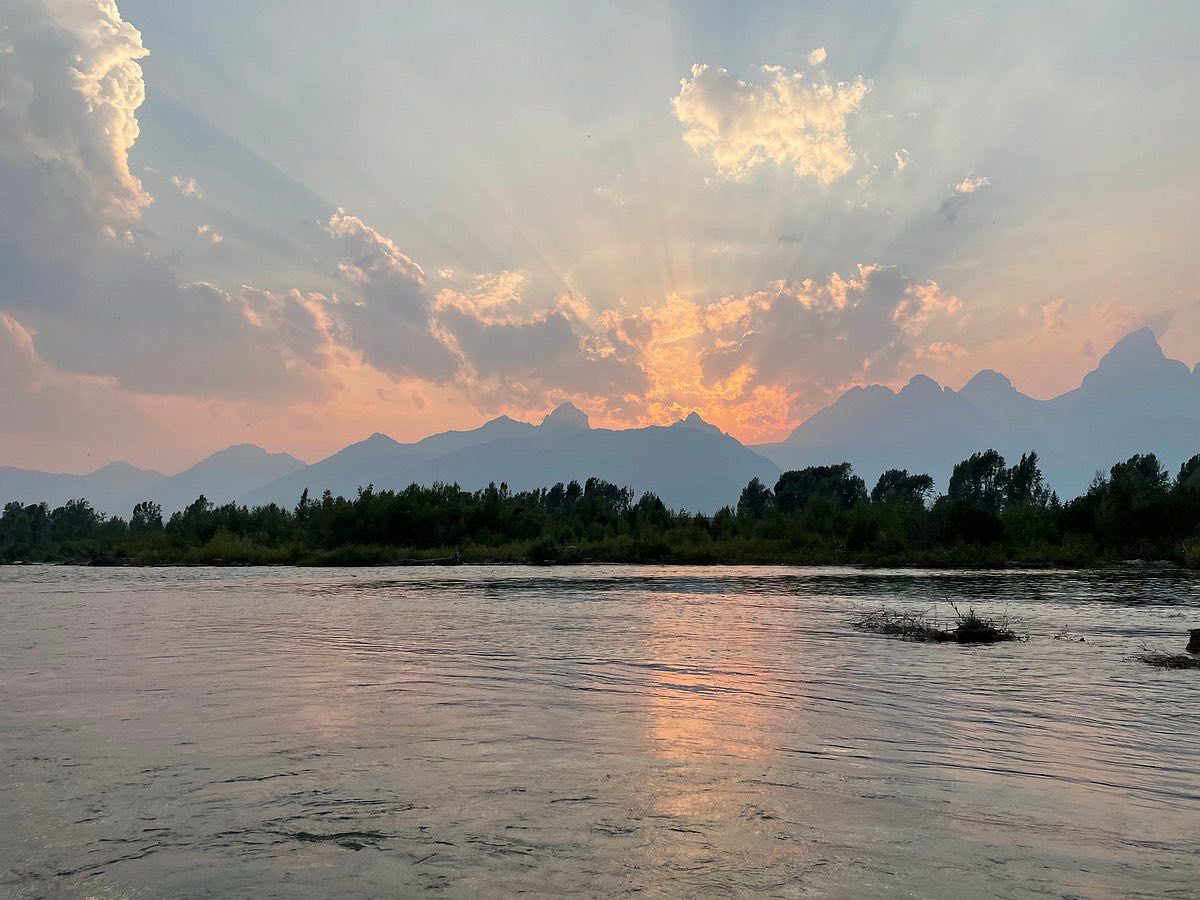 Sunset over a jagged mountain range with sunbeams radiating through clouds, silhouetted trees on a riverbank and pink-orange reflections on calm water