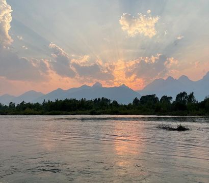 Sunset over a jagged mountain range with sunbeams radiating through clouds, silhouetted trees on a riverbank and pink-orange reflections on calm water