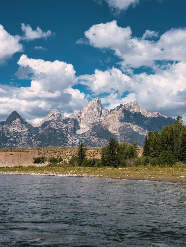Scenic river in the foreground with a rocky shore, pine trees and jagged mountain peaks under a bright blue sky dotted with fluffy white clouds.