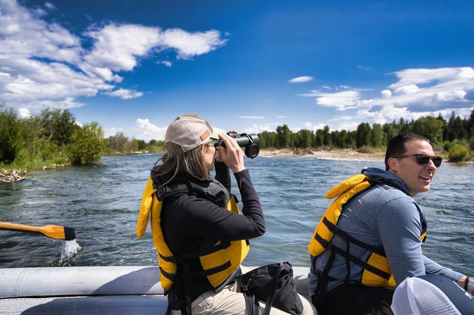 Two people in yellow life jackets on a raft on a scenic river, one scanning the tree-lined shore with binoculars while a paddle splashes under a bright blue sky — outdoor river adventure and wildlife viewing.