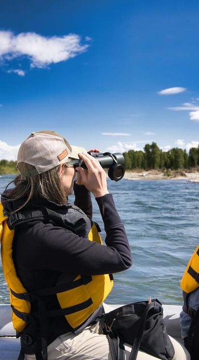 Two people in yellow life jackets on a raft on a scenic river, one scanning the tree-lined shore with binoculars while a paddle splashes under a bright blue sky — outdoor river adventure and wildlife viewing.