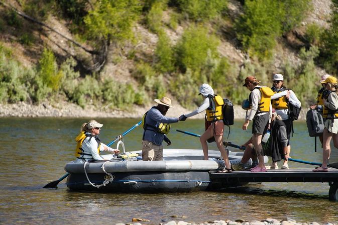 Group of rafters boarding an inflatable raft from a small dock on a sunny river, all wearing yellow life jackets and helping each other with oars against a forested, rocky shoreline — outdoor rafting trip adventure.