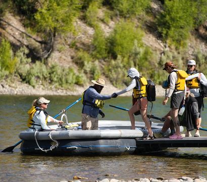 Group of rafters boarding an inflatable raft from a small dock on a sunny river, all wearing yellow life jackets and helping each other with oars against a forested, rocky shoreline — outdoor rafting trip adventure.
