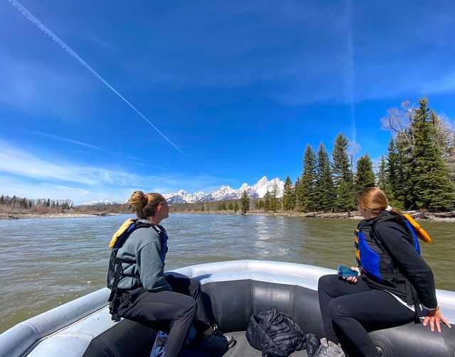 Two people in life jackets sitting in an inflatable raft on a wide mountain river, looking toward snow-capped peaks and a pine forest under a vivid blue sky with contrails.