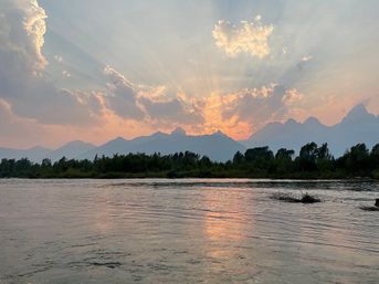 Serene river sunset with sun rays streaming through clouds over silhouetted mountains, tree-lined shore and orange-pink reflection on the water.