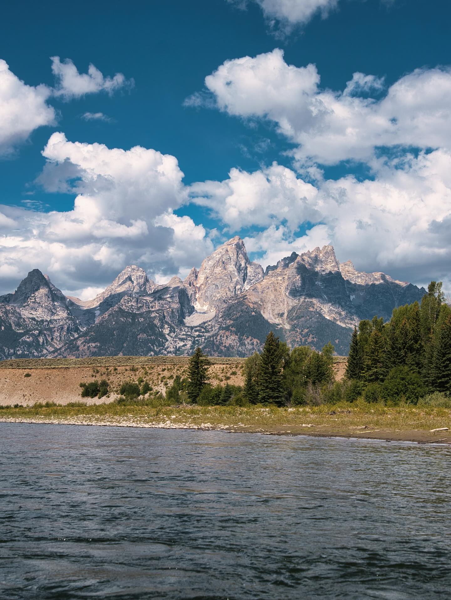 Calm river in the foreground with a pine-lined shore and jagged rocky mountains rising under a bright blue sky dotted with fluffy white clouds.