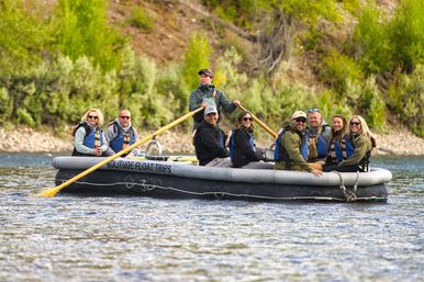 Guided river rafting trip: smiling group of adults on a large inflatable raft led by a standing guide rowing wooden oars on a calm river, passengers in life jackets with a green forested riverbank backdrop.