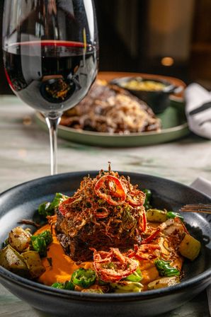 Gourmet seared steak topped with crispy fried shallots and sliced chilies on a creamy roasted pepper purée, served with roasted potatoes and a glass of red wine on a restaurant table.