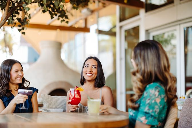 Three friends laughing and sipping colorful cocktails on a sunny outdoor restaurant patio with greenery and a wooden table, relaxed brunch vibe.