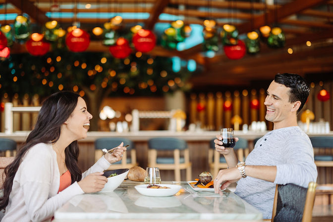 Smiling couple laughing over dinner at a stylish restaurant with colorful hanging lights; man lifts a glass of red wine while woman holds a salad bowl, plates and bread on the table.