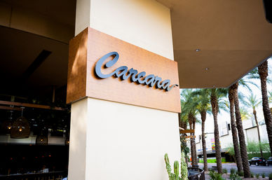 Cursive wooden restaurant sign mounted on a stucco column marking an open-air dining entrance beside a palm‑lined sidewalk