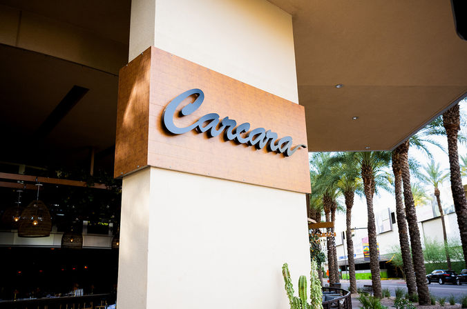 Cursive wooden restaurant sign mounted on a stucco column marking an open-air dining entrance beside a palm‑lined sidewalk