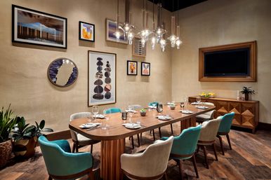 Modern private dining room interior with long walnut table set for eight, teal and beige upholstered chairs, sculptural glass pendant lights, framed gallery wall art, wooden sideboard and potted plants.