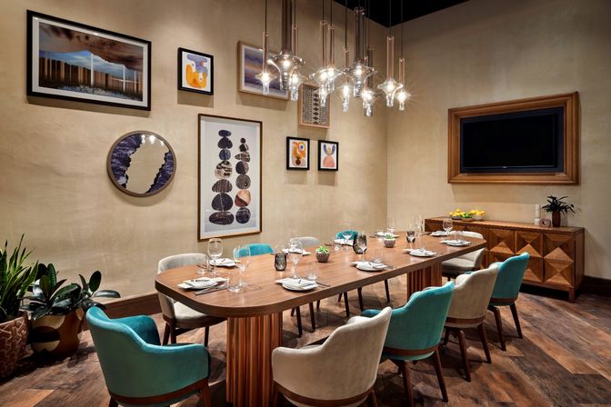 Modern private dining room interior with long walnut table set for eight, teal and beige upholstered chairs, sculptural glass pendant lights, framed gallery wall art, wooden sideboard and potted plants.