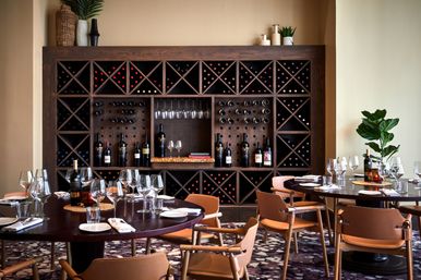 Stylish upscale restaurant dining room with a large wooden wine rack wall filled with bottles and hanging glasses, round tables set with wine glasses and plates, and tan leather chairs