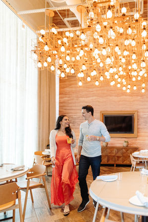 Smiling couple walking with cocktails through a modern hotel restaurant dining area beneath a large geometric chandelier of glowing bulbs, warm wood tables and round seating.