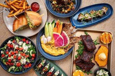 Overhead view of a colorful restaurant-style spread on a wooden table: grilled ribs with cornbread and charred orange, hummus with fresh veggies and watermelon-radish, watermelon-feta salad, sandwich with fries and ketchup, and assorted small bites.