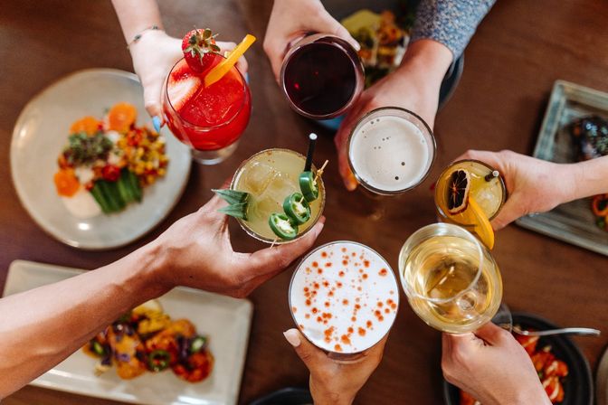 Top-down view of a group toast at a restaurant table with colorful drinks — strawberry cocktail, jalapeño margarita, beer, white wine and a foam-topped craft cocktail, surrounded by small plates of food.