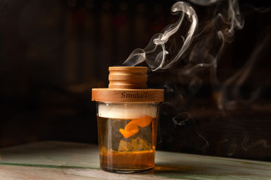 Smoked whiskey cocktail in a short glass with an orange peel, sealed under a wooden smoking lid as wisps of curled smoke rise in moody bar lighting.