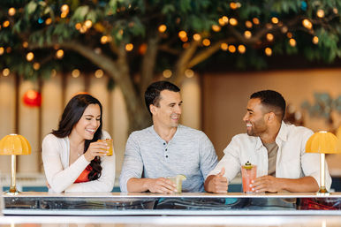 Three friends laughing at an indoor bar counter, sipping colorful cocktails beneath warm bokeh string lights and a decorative tree