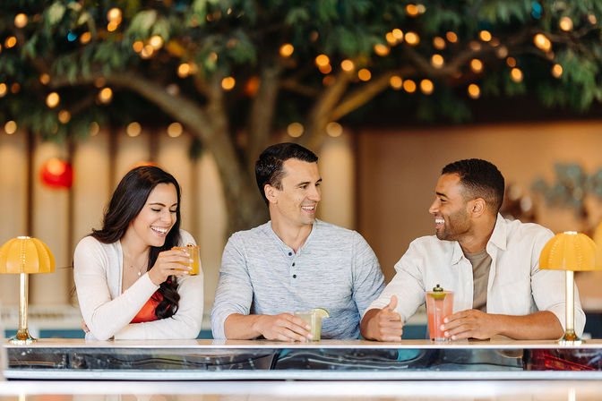 Three friends laughing at an indoor bar counter, sipping colorful cocktails beneath warm bokeh string lights and a decorative tree