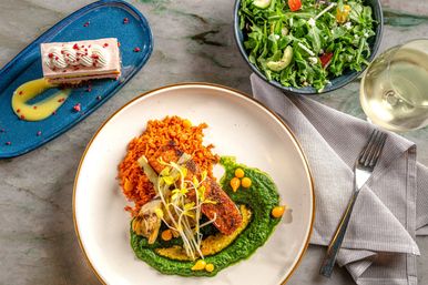 Top-down view of a gourmet seared salmon on bright green spinach purée with seasoned orange rice, microgreens and yellow sauce drops, paired with an arugula salad, layered dessert on a blue plate and a glass of white wine on a marble table.