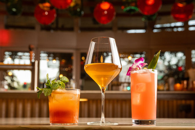 Three sunset-hued cocktails on a restaurant bar counter: a mint-garnished short orange drink, an amber wine in a stemmed glass, and a pink highball topped with an orchid, warm bokeh lights in the background.