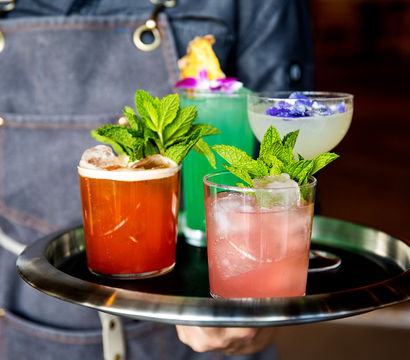 Server holding a tray with four colorful craft cocktails — pink, orange, turquoise and pale yellow — each topped with fresh mint and edible flowers at a cocktail bar.