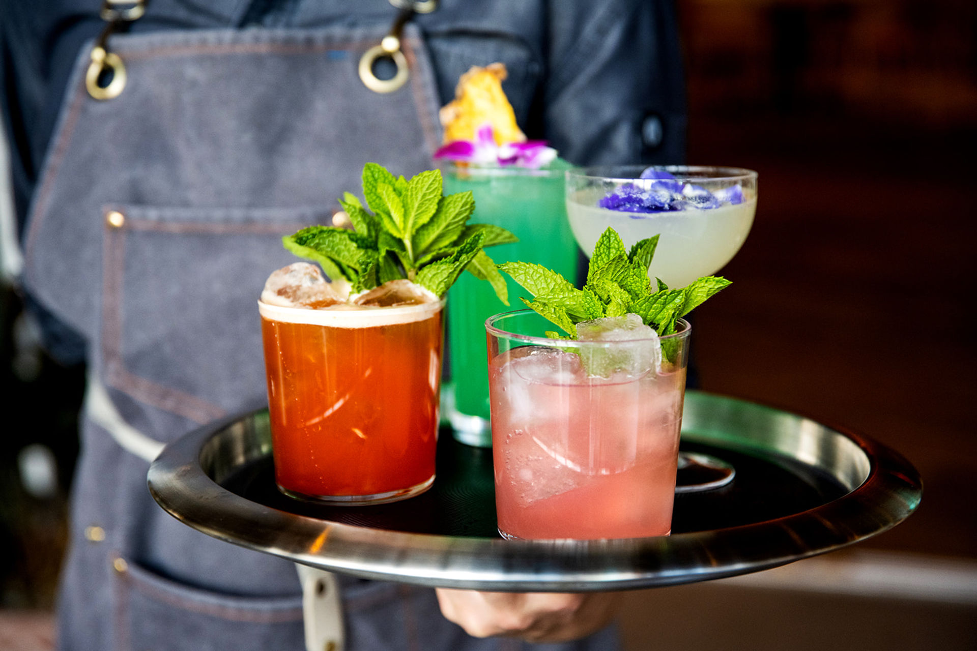 Server holding a tray with four colorful craft cocktails — pink, orange, turquoise and pale yellow — each topped with fresh mint and edible flowers at a cocktail bar.