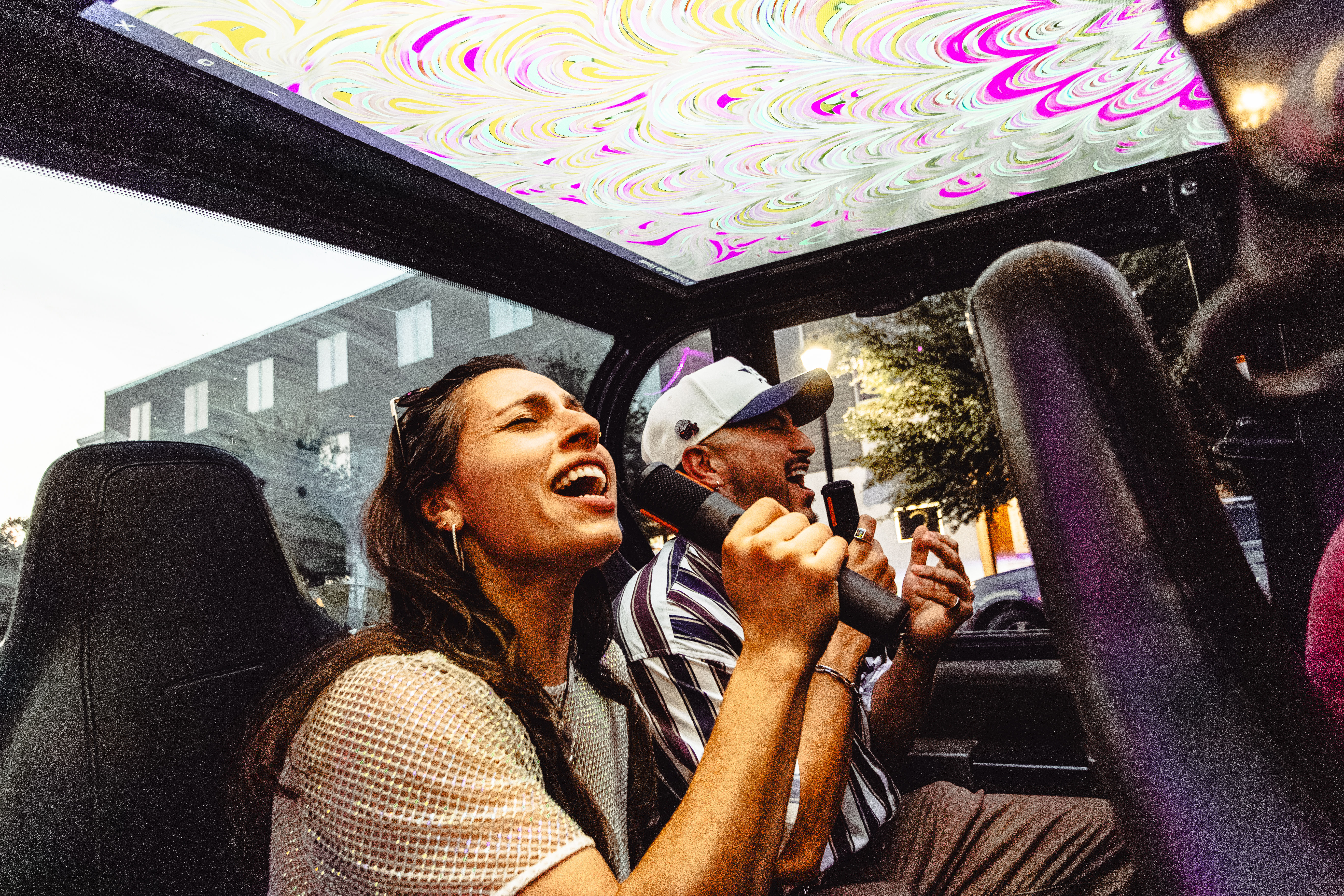 Two adults singing into handheld microphones inside a car with a colorful psychedelic sunroof, enjoying outdoor urban karaoke on a city street at dusk.