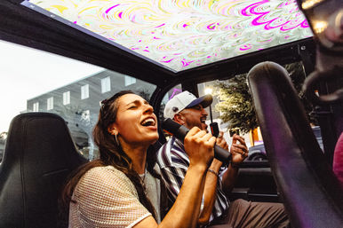 Two adults singing into handheld microphones inside a car with a colorful psychedelic sunroof, enjoying outdoor urban karaoke on a city street at dusk.