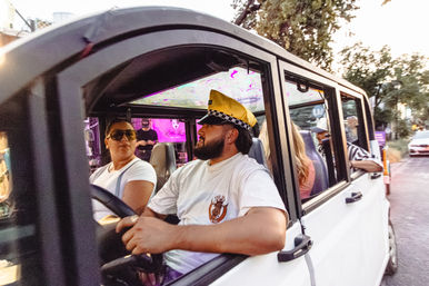Group of people riding in a white open-sided vehicle down a downtown street at dusk, driver wearing a yellow checkered taxi-style cap and passengers chatting under a colorful LED-lit ceiling