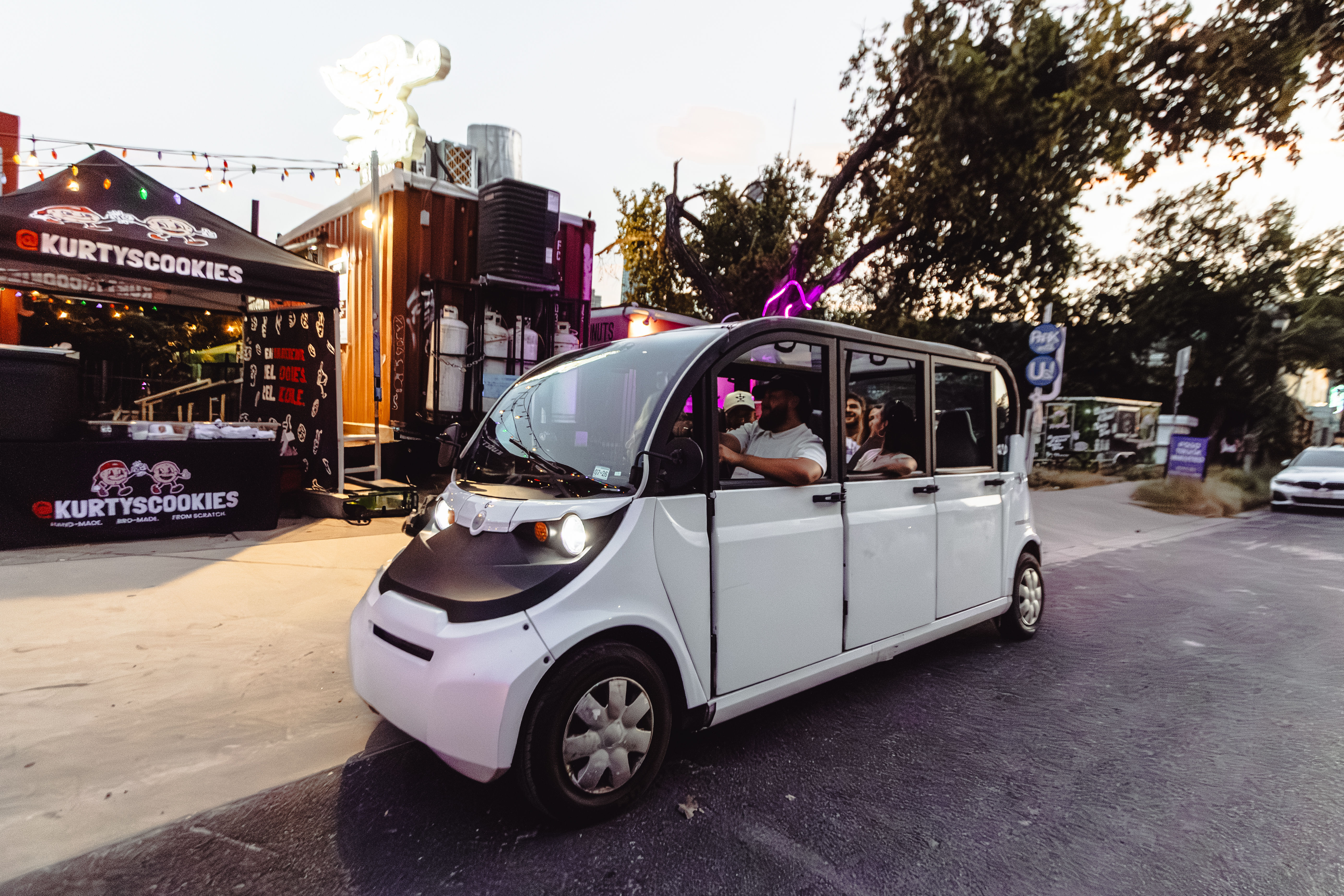 White electric shuttle carrying passengers past pop-up food stalls and neon-lit vendor tents on an urban street at dusk