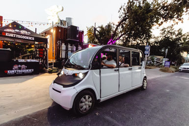 White electric shuttle carrying passengers past pop-up food stalls and neon-lit vendor tents on an urban street at dusk