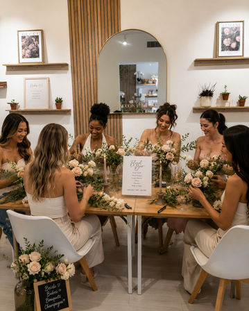 Six women laughing around a wooden table in a modern floral studio, creating blush rose bouquets at a DIY flower bar with greenery and champagne