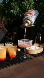 Hand pouring a pink cocktail from a metal-top shaker into a salt-rimmed coupe glass on a sunny backyard patio, with three orange-red layered drinks in plastic cups nearby.