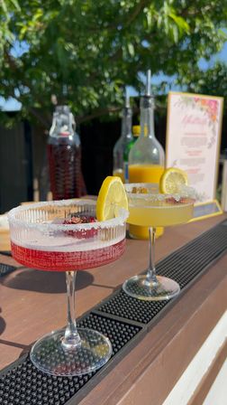 Sunlit outdoor bar with two sugar-rimmed coupe cocktails — a red berry drink and a yellow citrus drink each garnished with a lemon slice, bottles and menu blurred in the background for a summery patio vibe.