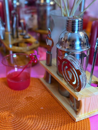 Stainless steel cocktail shaker and coiled strainer on a wooden bar-tool rack beside a pink cocktail in a plastic cup on an orange woven placemat — colorful home bar party setup.