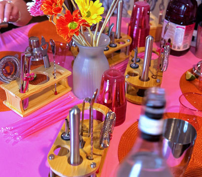 Bright gerbera daisies in a gray vase on a pink tablecloth surrounded by wooden cocktail-tool racks, stacked red party cups and bottles — colorful party bar centerpiece.