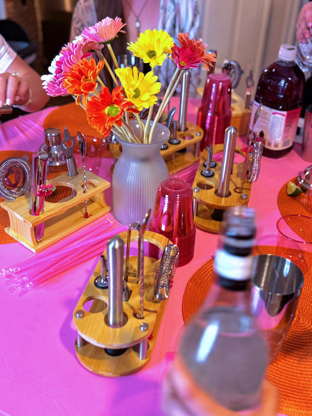 Bright gerbera daisies in a gray vase on a pink tablecloth surrounded by wooden cocktail-tool racks, stacked red party cups and bottles — colorful party bar centerpiece.